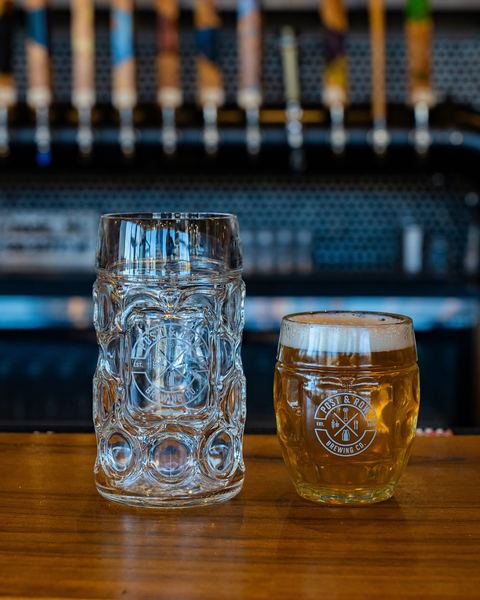An empty Festbier stein and a pint of golden lager, both etched with the round Post & Row Brewing Co. logo, on the bar in front of the taps