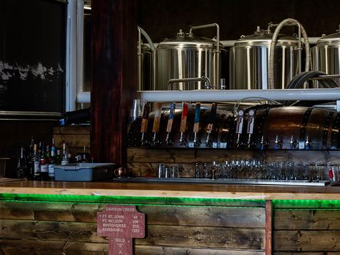 Our brewery bar: a row of tap handles in front of the stainless brewhouse kettles, with the Mile 0 signpost on the wooden bar front