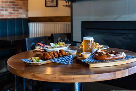 A round wooden table set with wings, a soft pretzel, poutine, chips and dip and a Post & Row Brewing Co. pint