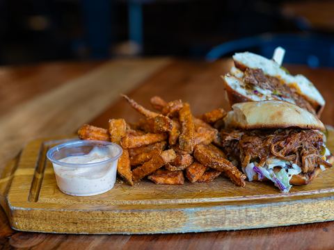 A BBQ pulled pork sandwich on a wooden board with yam fries and a creamy dip