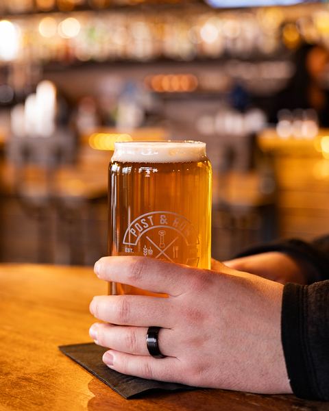 A hand holding a pint in a Post & Row Brewing Co. glass at the bar